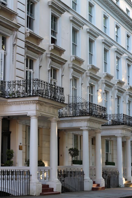 A multi-storey white residential building in Belgravia with ornate architectural details, including decorative cornices, large sash windows, and black wrought-iron balconies. The ground floor features a covered entrance area supported by white columns, with a small outdoor steps leading up to the door. In front of the building, there are pavement and a low black metal fence. The image captures the exterior during daylight with natural lighting, emphasizing the classic Georgian style. Belgravia Movers may be involved in the home relocation process for this property, possibly managing furniture transport or packing and moving activities around the building's front area.