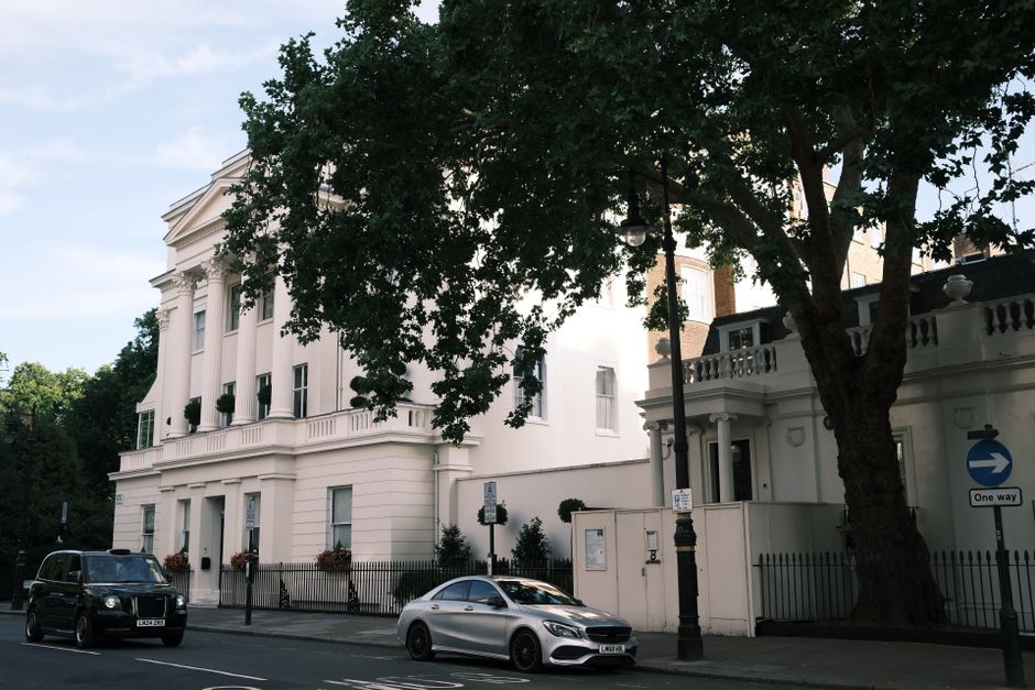 The image shows a street view of a large, elegant white building with classical architectural features, including tall windows and decorative columns, situated in Belgravia near Eaton Square Mansions. In front of the building, there are two parked vehicles: a black van and a silver sedan, both positioned parallel to the pavement. A black metal fence runs along the front of the property, with potted plants and small shrubs visible inside the enclosed area. A large mature tree with dense foliage partially obscures part of the building and provides shade, with sunlight filtering through the leaves. A vintage-style black street lamp stands beside the sidewalk, which features a one-way traffic sign indicating the direction of travel. The environment suggests a quiet, affluent neighbourhood suitable for professional house removals or furniture transport services. This scene could depict the setting for a home relocation process, with the façade of a period property ready for packing and moving activities, as performed by companies such as Belgravia Movers.