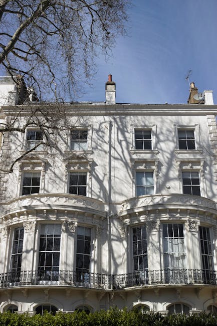 A multi-storey white residential building in Belgravia with ornate architectural details, including decorative cornices, large sash windows, and black wrought-iron balconies. The ground floor features a covered entrance area supported by white columns, with a small outdoor steps leading up to the door. In front of the building, there are pavement and a low black metal fence. The image captures the exterior during daylight with natural lighting, emphasizing the classic Georgian style. Belgravia Movers may be involved in the home relocation process for this property, possibly managing furniture transport or packing and moving activities around the building's front area.