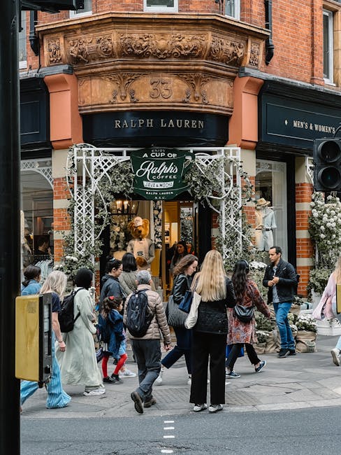 A busy street scene outside a Ralph Lauren store located at 53 Sloane Street, with a decorative floral arch overhead and a sign for Ralph's Coffee hanging beneath. The storefront features large windows displaying mannequins dressed in retail clothing, and the building has ornate brickwork and detailed architectural elements above the entrance. In front of the shop, numerous pedestrians are walking and standing on the pavement, including people carrying shopping bags and children, all dressed in casual attire. The scene captures a typical day of shopping and foot traffic in the area, with some individuals engaged in conversation and others looking at the store's window displays. The lighting suggests it is daytime, likely in overcast weather, adding a neutral and realistic tone to the scene, while the overall environment reflects a vibrant shopping district, ideal for house removals and relocation services like those offered by Belgravia Movers.