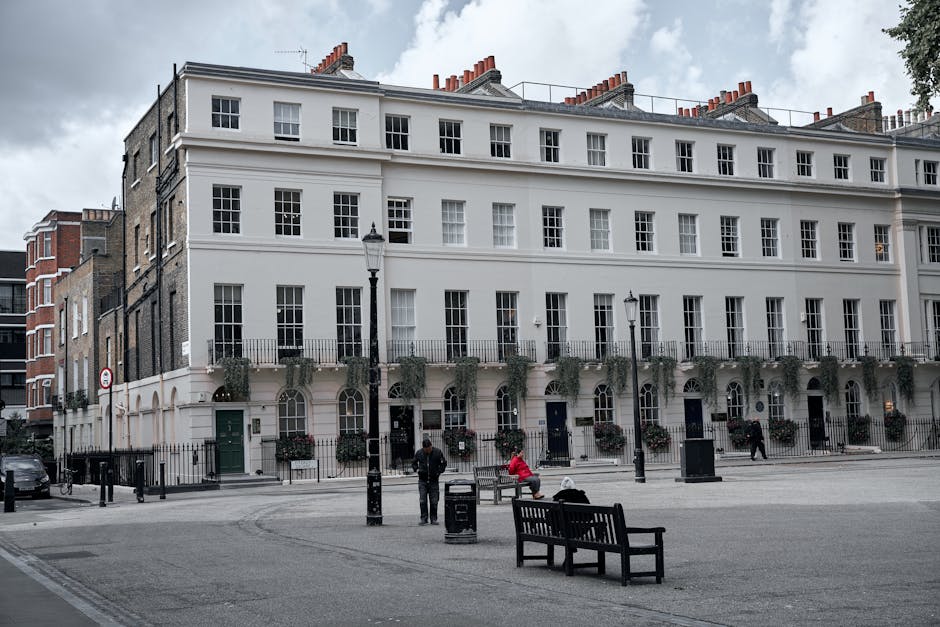 A wide-angle view of a street scene outside a white multi-storey residential building with numerous uniformly sized windows and black iron railings, typical of a London townhouse. In the foreground, t