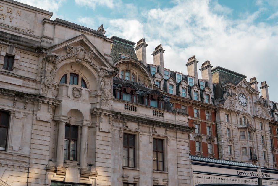 A black and white photograph of a narrow urban street lined with multi-storey Victorian buildings featuring ornate facades, decorative window surrounds, and sloped roofs with dormer windows. Parked ca